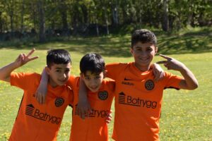Tre glada killar med mörkt hör och orange fotbollströjor håller om varandra och ler in i kameran.